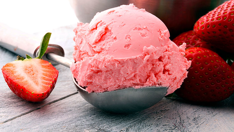 Eine Kugel Erdbeereis in einer Eiskelle vor frischen Erdbeeren und einer halbierten Erdbeere auf grauem Holzuntergrund.
