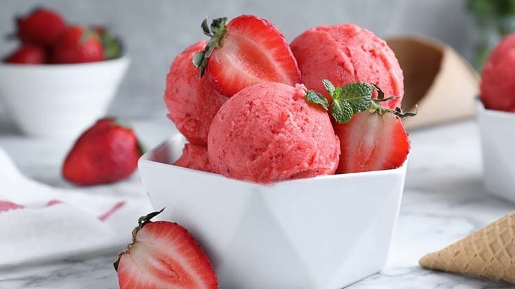 Rosa Erdbeereis mit frischen Erdbeeren und Minzblättern in einer weißen Schale, daneben liegt eine leere Eiswaffel.