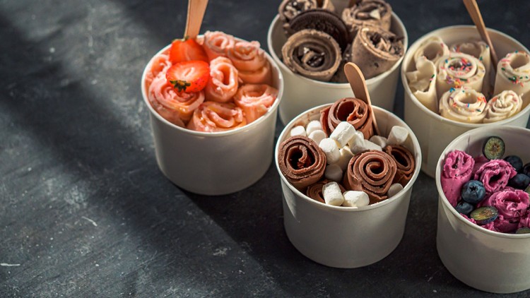 Fünf Becher mit gerolltem Eis in verschiedenen Farben und Dekorationen wie Früchten, Keksen, Marshmallows und Streuseln.
