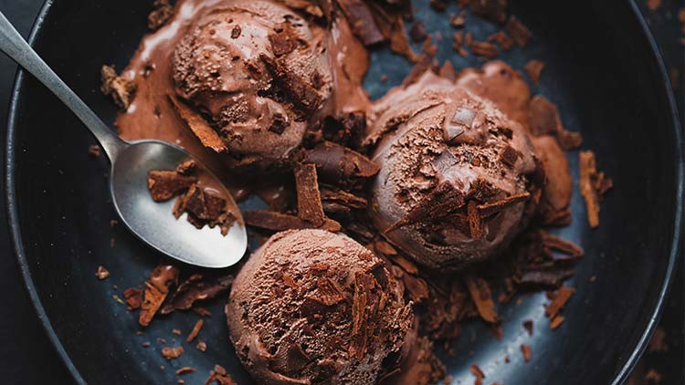 Drei Kugeln Schokoladeneis mit Schokoraspeln in einer schwarzen Schale, daneben ein Löffel mit Schokostückchen.