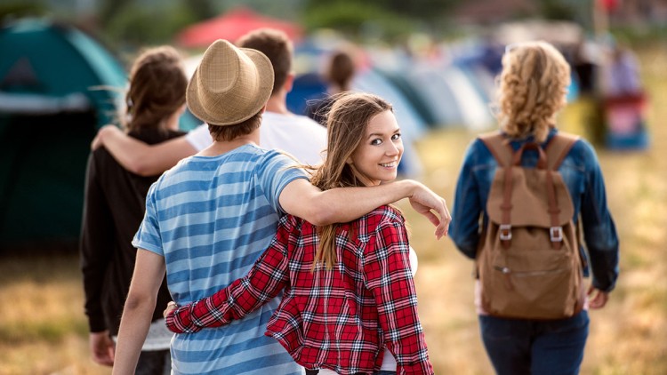 Mehrere Personen gehen auf einer Wiese mit Zelten im Hintergrund, eine Person trägt einen Strohhut und eine gestreifte Bluse.