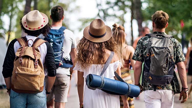 Eine Gruppe von vier Personen geht mit Rucksäcken und einer gerollten Matte auf einem Weg durch einen grünen Park.