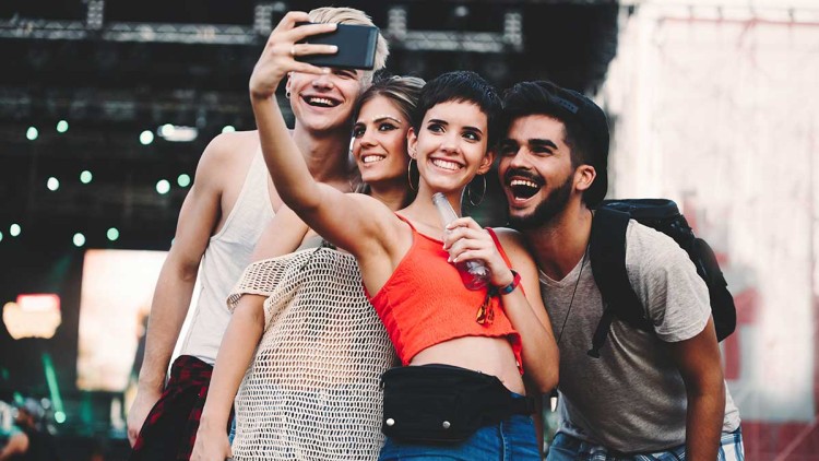 Vier Personen stehen dicht beieinander vor einer Konzertbühne und machen ein Selfie, im Hintergrund sind Lichter und Menschen.