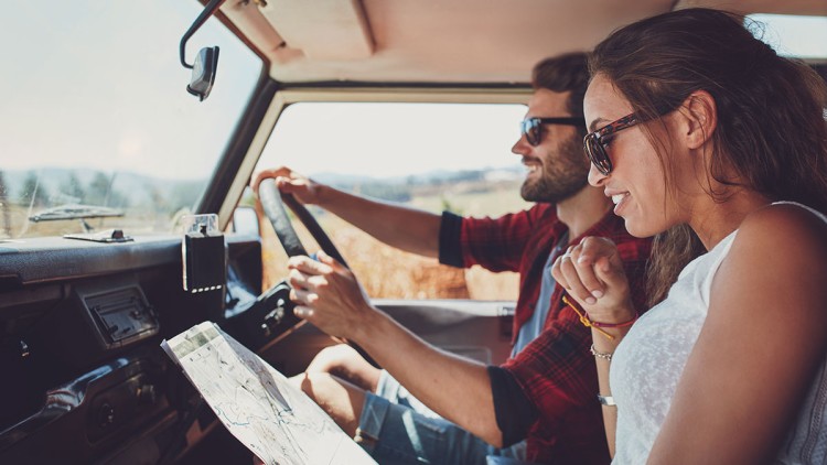 Person am Steuer eines Autos im Innenraum, andere Person hält eine Landkarte, im Hintergrund ländliche Landschaft.