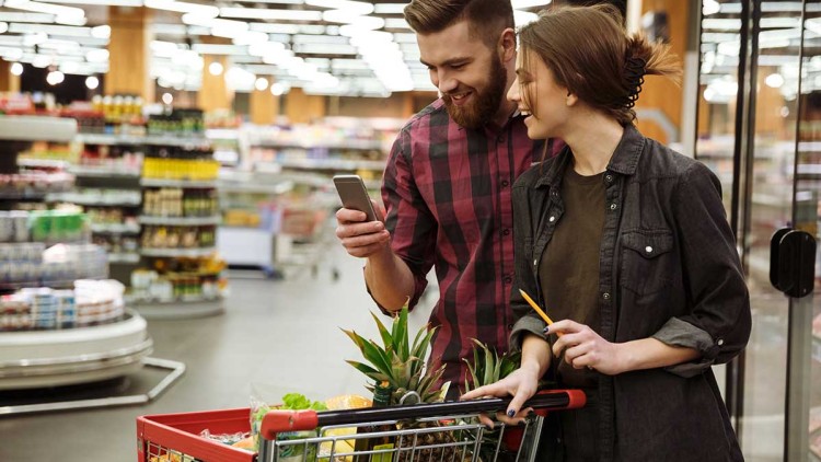 Ein Einkaufswagen mit Obst und Gemüse wird in einem modern beleuchteten Supermarkt geschoben, eine Person hält ein Smartphone.