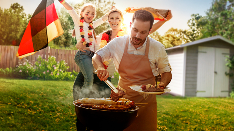 Person grillt Fleisch und Maiskolben im Garten, während im Hintergrund eine Person mit Deutschlandfahne feiert.