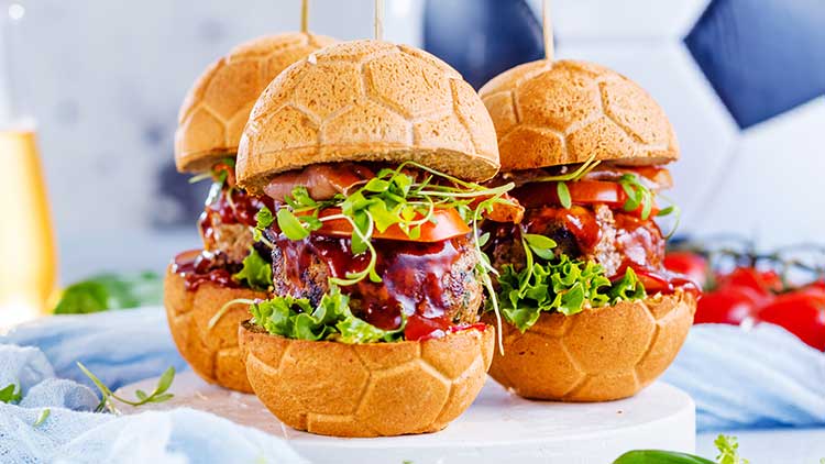 Drei Burger mit Brötchen in Fußballoptik, Salat, Tomaten und Sauce stehen auf einem Holzbrett, im Hintergrund ein Fußball.