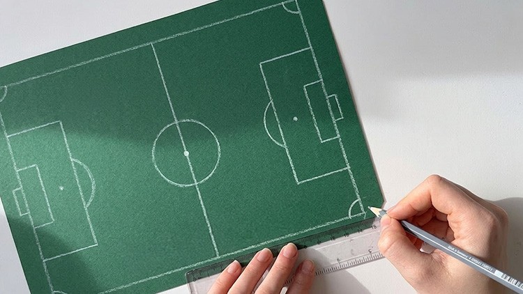 Grüne Karte mit weißem Fußballfeld, eine Hand zeichnet mit Bleistift und Lineal Linien am unteren Rand.