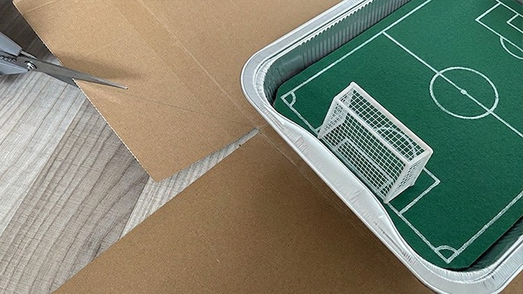 Grüne Spielfeldmatte mit Mini-Tor in einer großen Aluminiumform, daneben Karton und eine Schere.