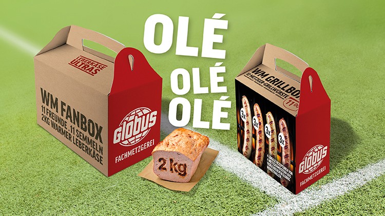 Zwei Kartons der GLOBUS Fachmetzgerei mit warmem Leberkäse und Grillwürstchen, davor ein 2 kg Leberkäse auf Papier.