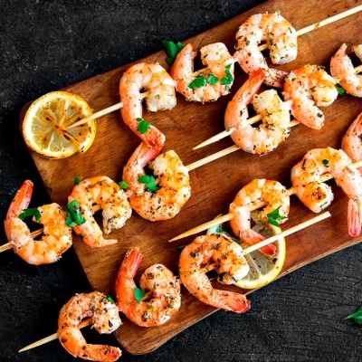 Gegrillte Garnelen an Holzspießen mit Kräutern, Zitronenscheiben und halbierter Knoblauchknolle auf einem Holzbrett.