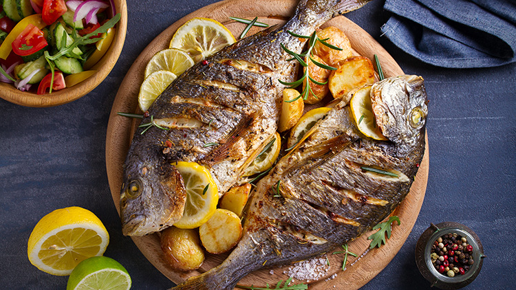 Zwei gegrillte Fische mit Zitronenscheiben und Rosmarin auf Ofenkartoffeln, daneben eine Schale mit Salat und halbe Zitrone.