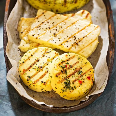 Gegrillter Käse in verschiedenen Formen mit braunen Grillstreifen und Kräutern auf Backpapier in einer dunklen Schale.