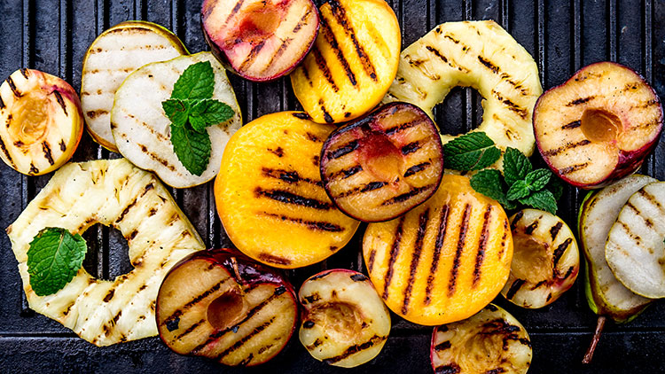 Gegrillte Scheiben von Pfirsichen, Ananas, Birnen und Pflaumen mit Minzblättern auf einem schwarzen Grillrost.
