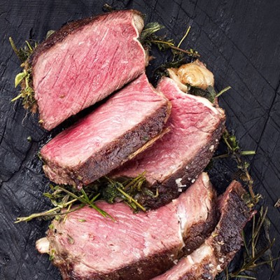 Fünf saftige, rosa gebratene Steakstücke liegen auf einer dunklen Holzunterlage, dekoriert mit Kräutern und grobem Salz.