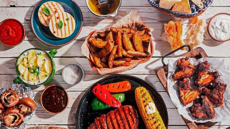 Gegrilltes Fleisch, bunte Paprika, Maiskolben, Kartoffelspalten, Brot, Saucen, Pilze, Salat und Würstchen auf Holztisch.