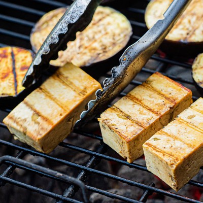 Tofustücke und Auberginenscheiben mit Grillstreifen auf einem Rost, eine Grillzange hält ein Tofustück fest.