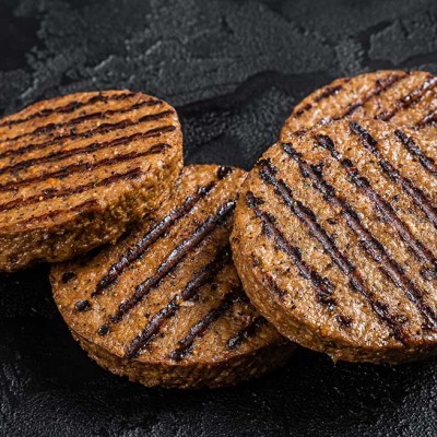Vier gegrillte Burger-Patties mit Grillstreifen liegen auf einer dunklen, strukturierten Oberfläche.