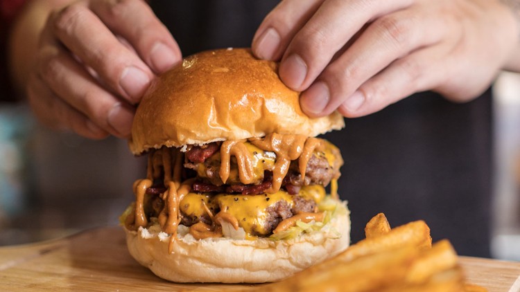 Hände halten einen Burger mit Käse, Soße, Bacon und Salat, daneben liegen Pommes auf einem Holzbrett.