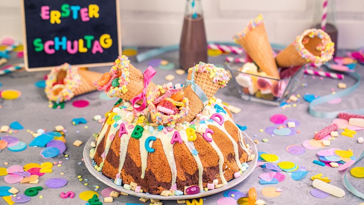 Bunter Marmorkuchen mit Zuckerguss, Buchstaben-Deko und gefüllten Waffeltüten, daneben Tafel mit „ERSTER SCHULTAG“.