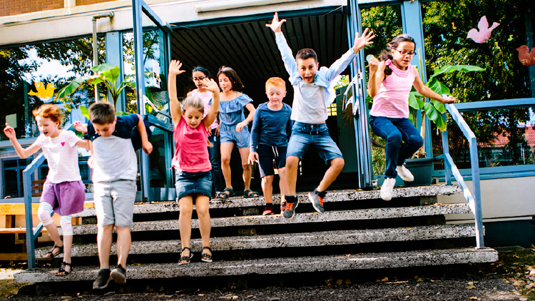 Gruppe von Kindern springt freudig eine Treppe vor einem Gebäude mit großen Fenstern und Pflanzen im Hintergrund hinunter.
