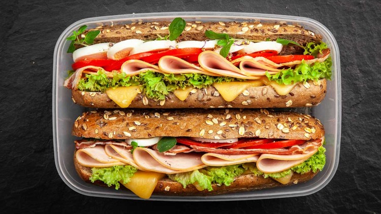 Zwei Mehrkornbrötchen in einer Plastikbox, belegt mit Käse, Salat, Schinken, Tomaten, Ei und Kräutern.