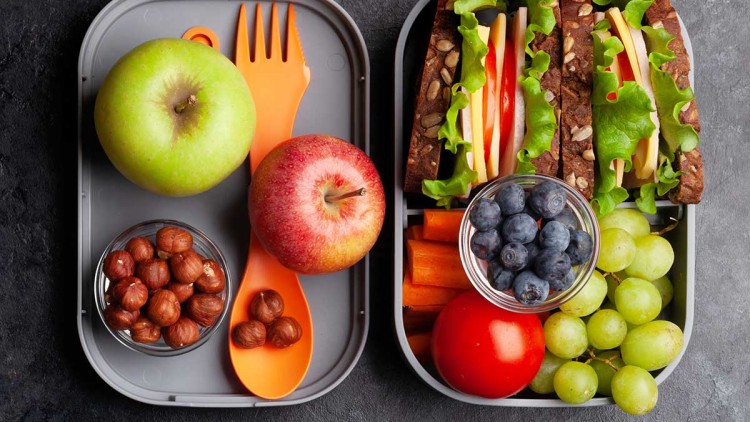 Zwei graue Lunchboxen mit Obst, Gemüse, Nüssen, einem orangefarbenen Besteck und zwei belegten Vollkornbrotscheiben.