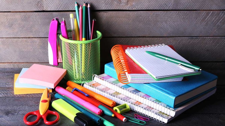 Schul- und Büromaterialien wie Hefte, Stifte, Scheren, Heftklammern und Klebezettel liegen auf einem Holztisch.