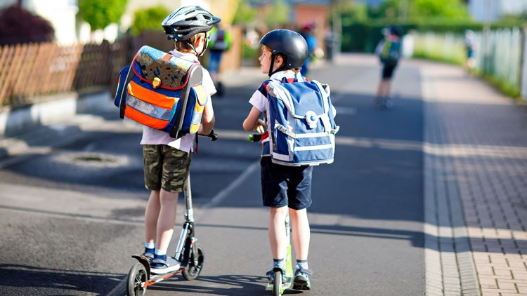 Zwei Kinder mit Schulranzen und Helmen fahren auf Rollern eine sonnige Wohnstraße entlang.