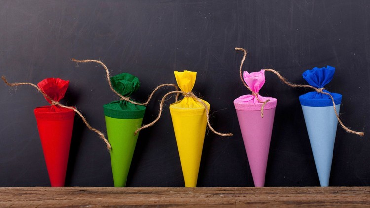 Fünf bunte Schultüten in Rot, Grün, Gelb, Rosa und Blau stehen vor einer dunklen Tafel auf einem Holztisch.
