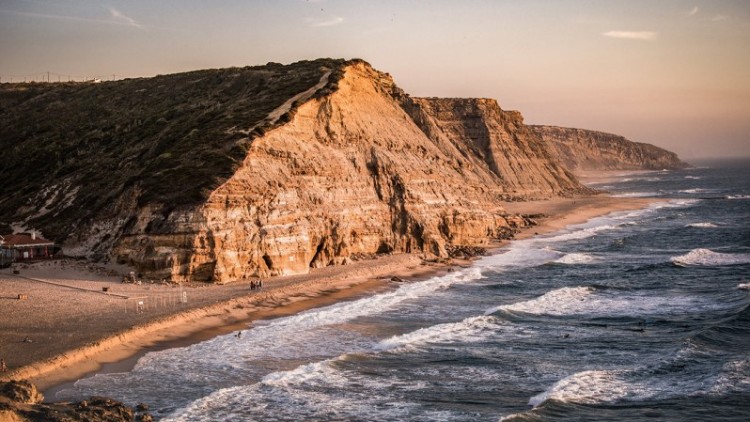 Steile Küstenklippen aus Sandstein grenzen an einen Strand, mit brechenden Wellen und einem Gebäude links im Bild.