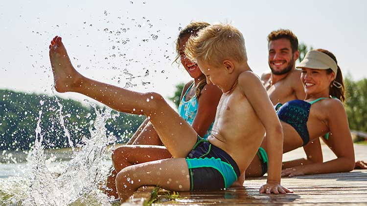 Menschen sitzen auf einem Steg am Wasser, ein Kind im Vordergrund spritzt mit einem ausgestreckten Bein Wasserfontänen hoch.