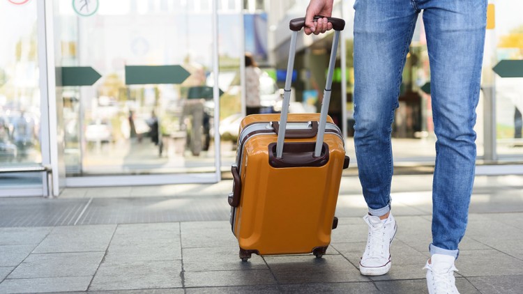 Person in blauer Jeans und weißen Schuhen zieht einen gelben Rollkoffer vor Glastüren mit Symbolen für Eingang und Ausgang.