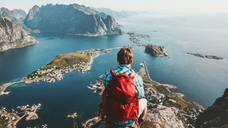 Person mit rotem Rucksack sitzt auf einem Felsen und blickt auf ein Dorf, Wasser und Berge in einer Küstenlandschaft.