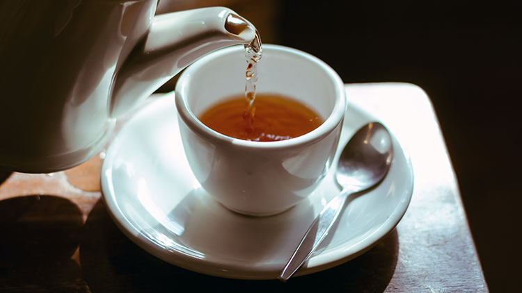 Tee wird aus einer weißen Kanne in eine weiße Tasse auf einer Untertasse mit Löffel gegossen.