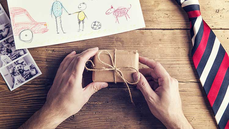 Hände mit kleinem Geschenk in braunem Papier und Schnur, daneben Kinderzeichnung, Fotos und gestreifte Krawatte auf Holz.