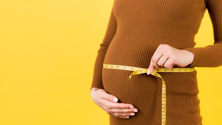 Person in braunem Strickkleid misst mit einem gelben Maßband den Bauchumfang, die linke Hand stützt den Bauch.