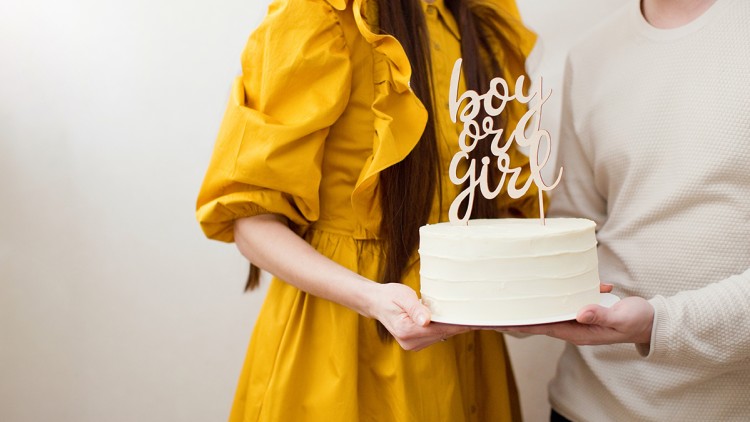 Zwei Personen halten eine weiße Torte mit der Aufschrift boy or girl auf einem Holz-Dekostäbchen.