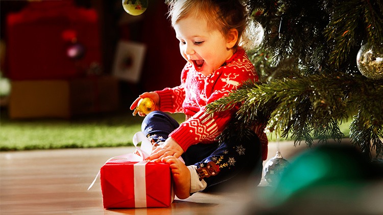 Kind in rotem Pullover sitzt auf dem Boden und hält mit einer Hand ein verpacktes Geschenk unter einem geschmückten Weihnachtsbaum.