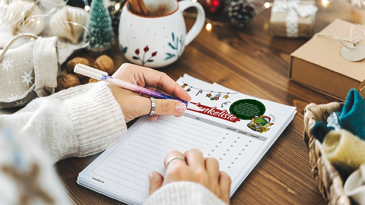 Hand schreibt mit einem lilafarbenen Stift auf eine Wunschliste, daneben Dekoration, Geschenke, Tasse und Korb mit Stoffen.