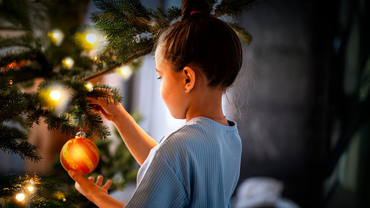 Kind dekoriert einen festlich beleuchteten Weihnachtsbaum mit einer roten Kugel, Hintergrund ist dunkel.