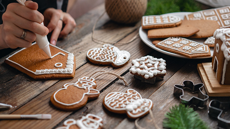Eine Hand verziert ein Lebkuchenhaus mit weißem Zuckerguss, während dekorierte Lebkuchen auf einem Holztisch liegen.