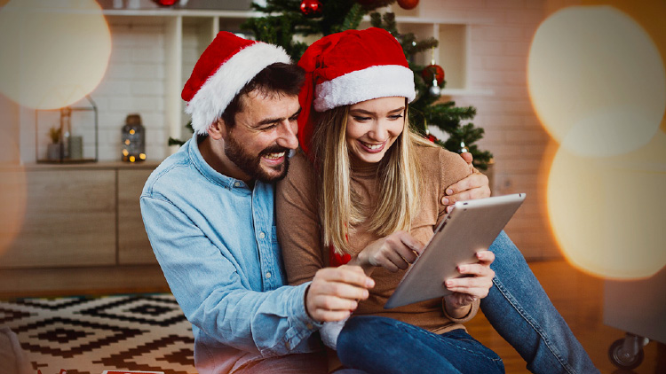 Zwei Personen mit Weihnachtsmützen sitzen vor einem geschmückten Tannenbaum und nutzen gemeinsam ein Tablet.
