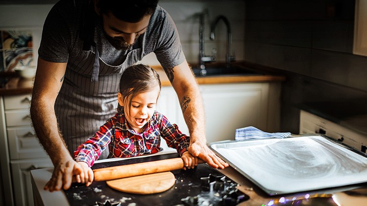 Ein Kind rollt mit einem Nudelholz Teig aus, während eine erwachsene Person unterstützend die Hände auf das Nudelholz legt.