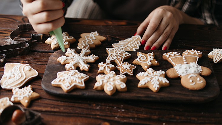 Hände verzieren mit grüner Spritztülle Lebkuchen in Stern- und Figurenform auf einem dunklen Holzbrett mit weißer Glasur.