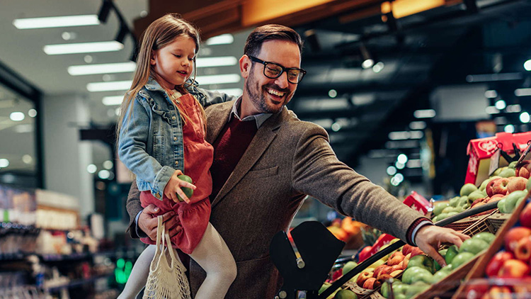 Eine Person im Anzug hält ein Kind auf dem Arm und greift nach Obst in einer Obst- und Gemüseabteilung eines Geschäfts.