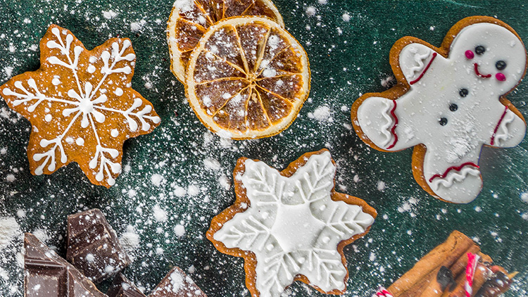 Lebkuchenmännchen, Schneeflockenplätzchen, Zimtsterne, Schokolade, Orangen und ein Nudelholz auf bemehltem Untergrund.