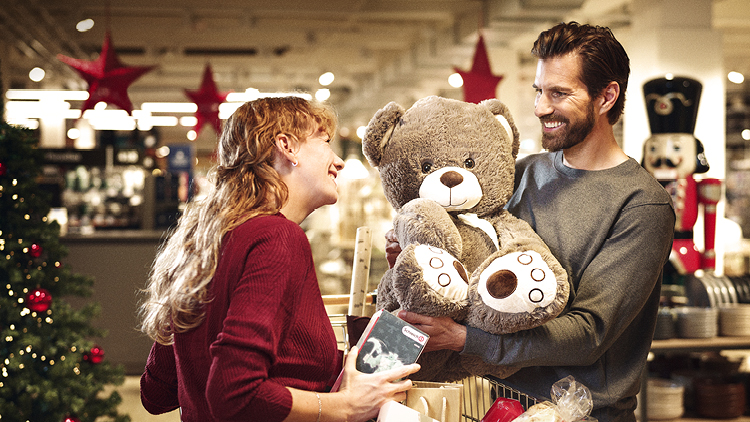 Eine Person hält einen großen, grauen Teddybären, im Hintergrund weihnachtliche Dekoration und geschmückte Tannenbäume.