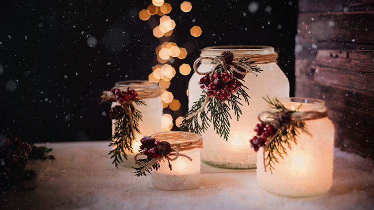 Vier gläserne Windlichter mit Kerzen, dekoriert mit Tannenzweigen, Beeren und Juteschnur, stehen auf Schnee vor Lichterketten.