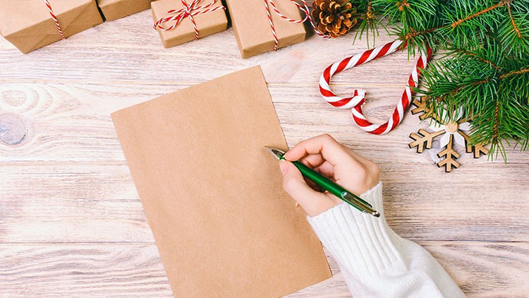 Ein grüner Kugelschreiber schreibt auf braunes Papier, daneben Geschenke, Tannenzweige, Zuckerstangen und ein Schneeflockenanhänger.